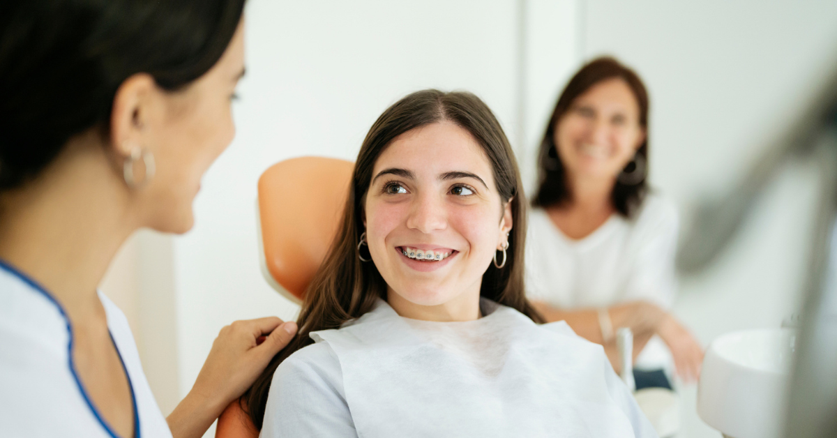 A teenage girl with braces sits in a dentist's chair, smiling at a female dentist. A woman in the background smiles warmly, creating a cheerful atmosphere.