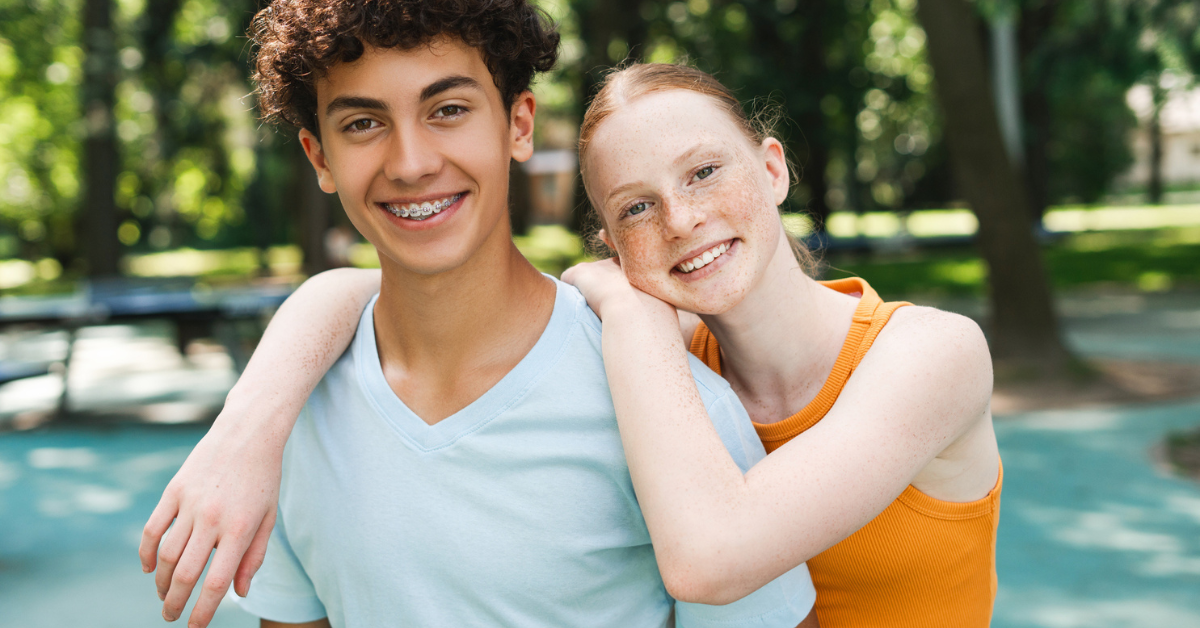 Does insurance cover braces? Two smiling teenagers, one with curly hair and braces, the other with freckles, embrace in a sunny park. The scene conveys happiness and friendship.