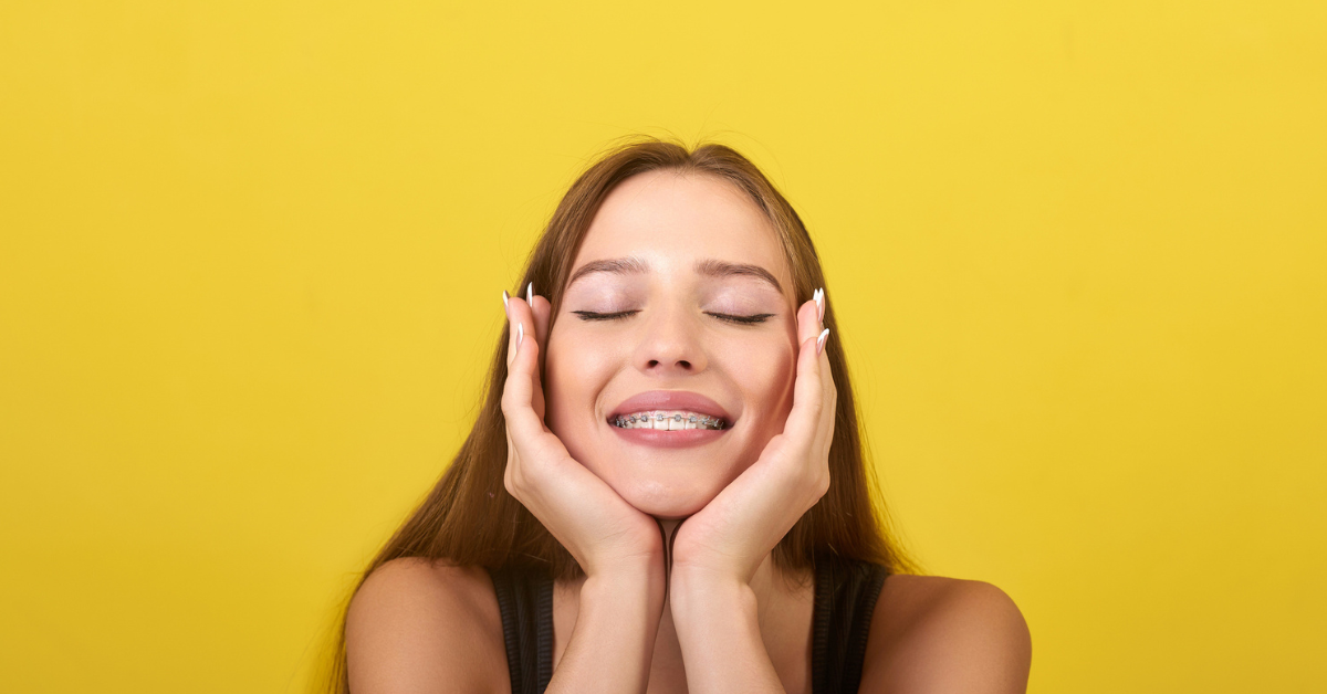 Orthodontics for teens - A young woman with long brown hair and braces smiles with closed eyes, gently cupping her face with her hands against a bright yellow background.