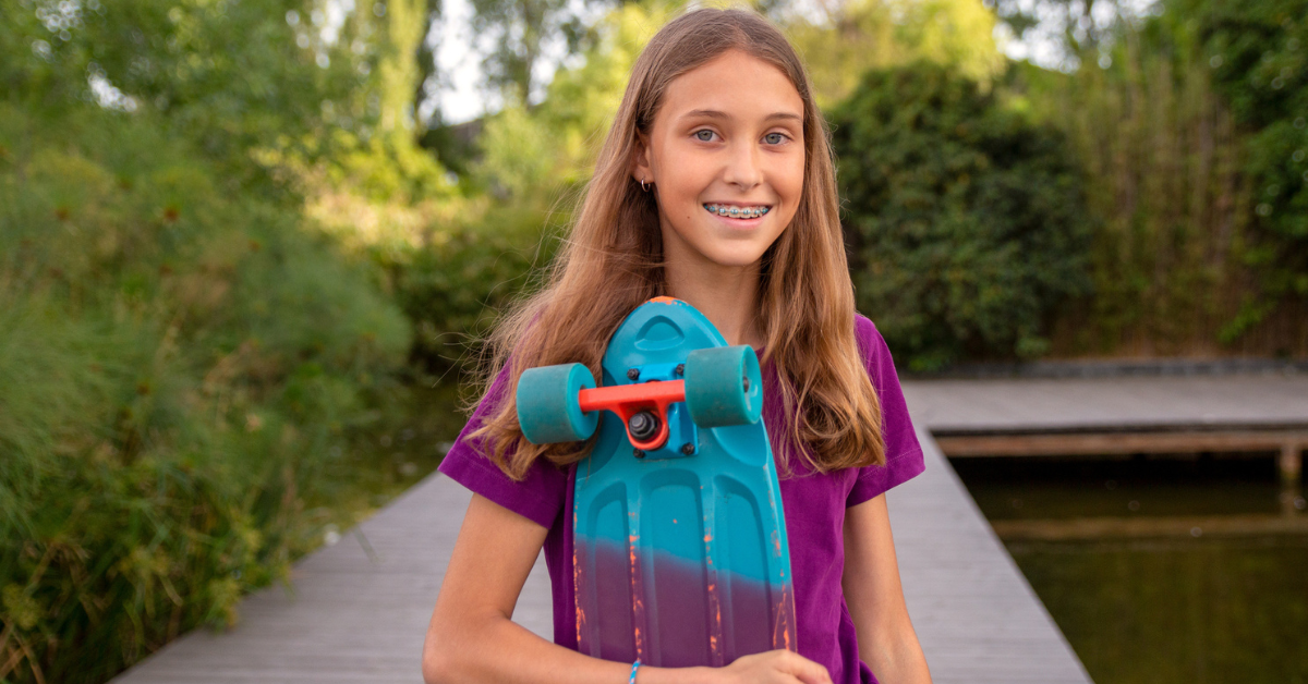 Smiling girl with braces holds a turquoise skateboard by a pond. Wearing a purple shirt, she stands on a wooden path surrounded by greenery.