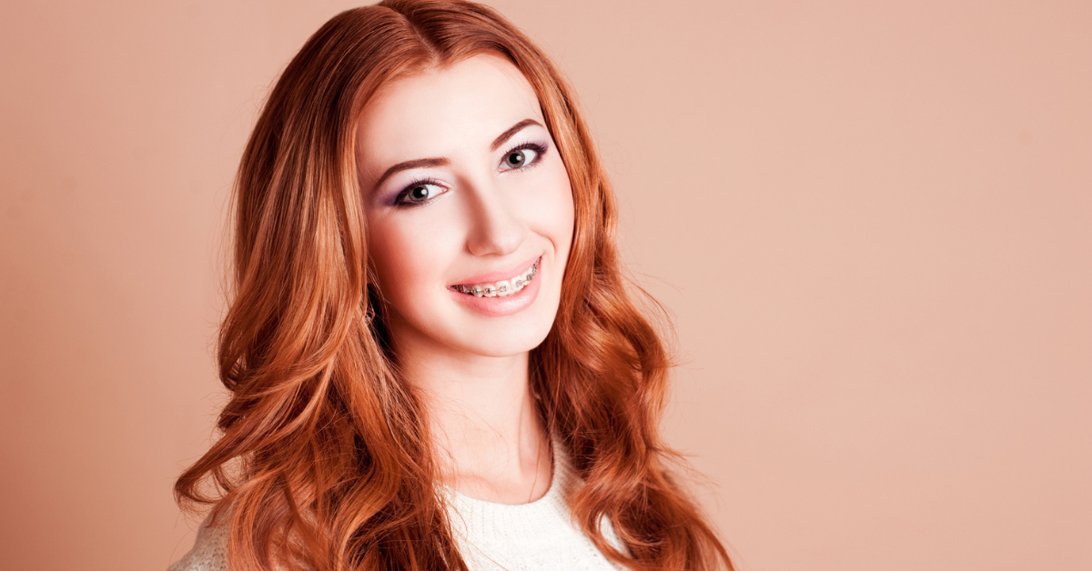 A smiling young woman with long auburn hair and braces on her teeth looks confidently at the camera. She wears a white sweater, set against a beige background.