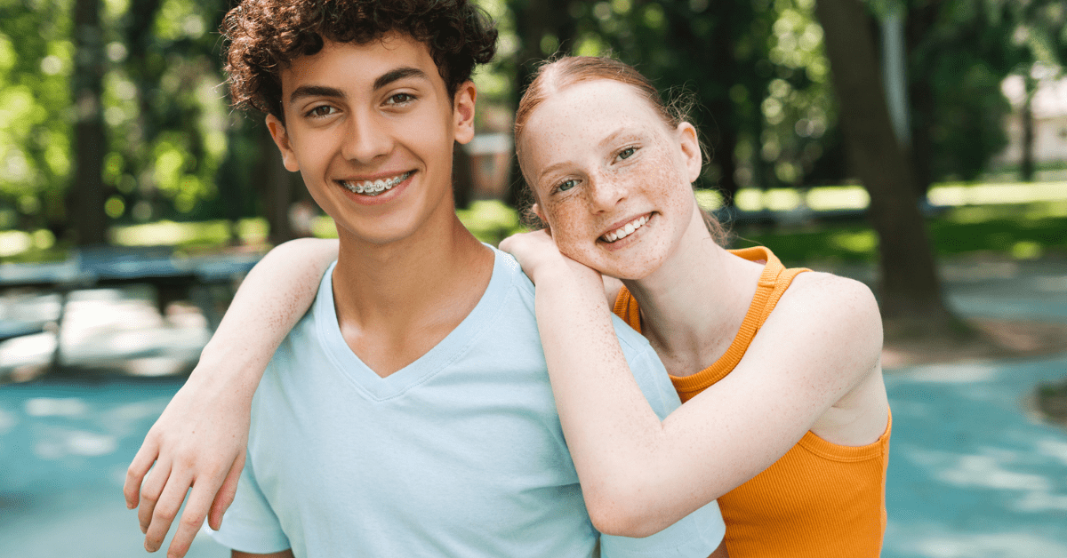 Does insurance cover braces? Two smiling teenagers, one with curly hair and braces, the other with freckles, embrace in a sunny park. The scene conveys happiness and friendship.