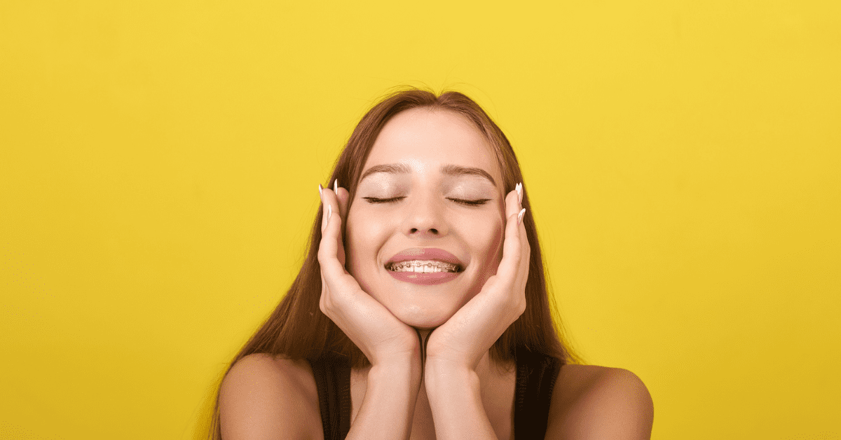Orthodontics for teens - A young woman with long brown hair and braces smiles with closed eyes, gently cupping her face with her hands against a bright yellow background.