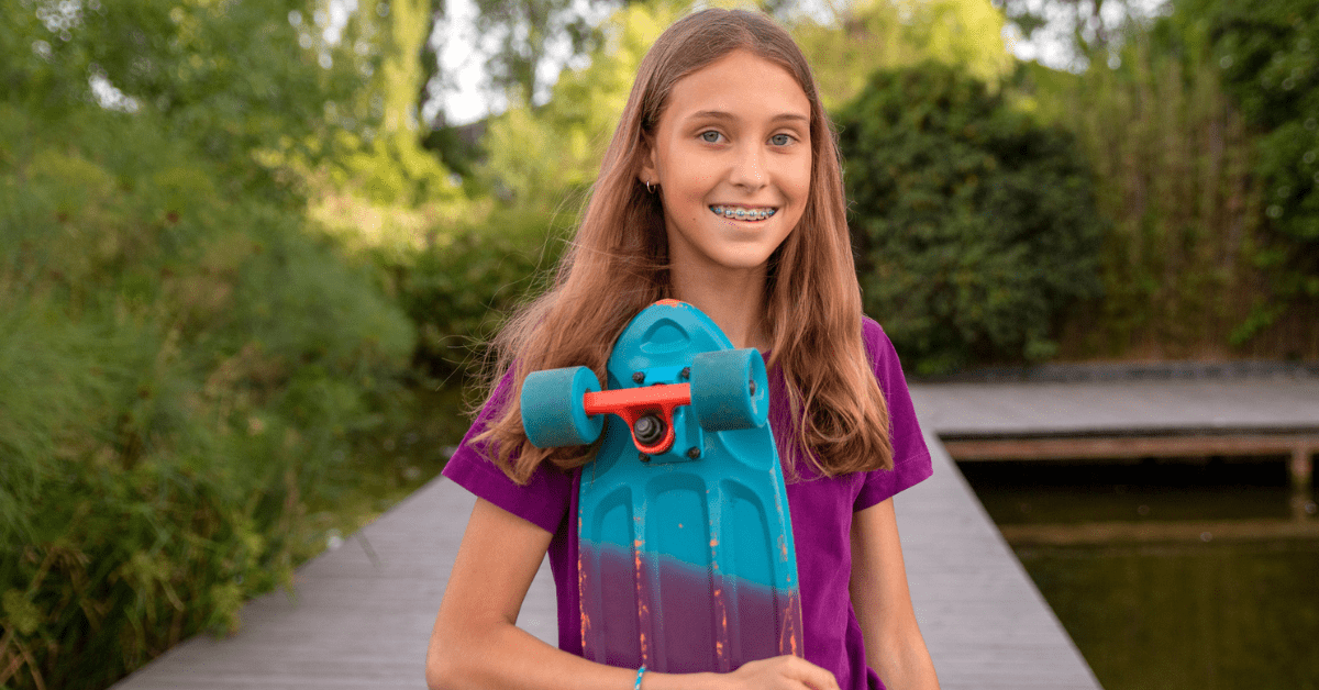 Smiling girl with braces holds a turquoise skateboard by a pond. Wearing a purple shirt, she stands on a wooden path surrounded by greenery.